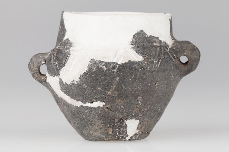 Waza czerniona z wysoką szyjką, baniastym brzuścem i płaskim dnem. Naczynie ornamentowane dookolną linią rytą.