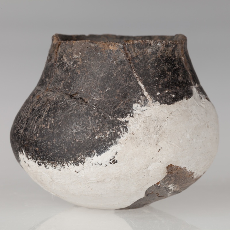 Miniaturowa waza o płaskim dnie, brzuścu baniastym i stożkowatej szyjce. Naczynie zdobione dookolnym żłobkiem i naprzemiennymi grupami kresek.