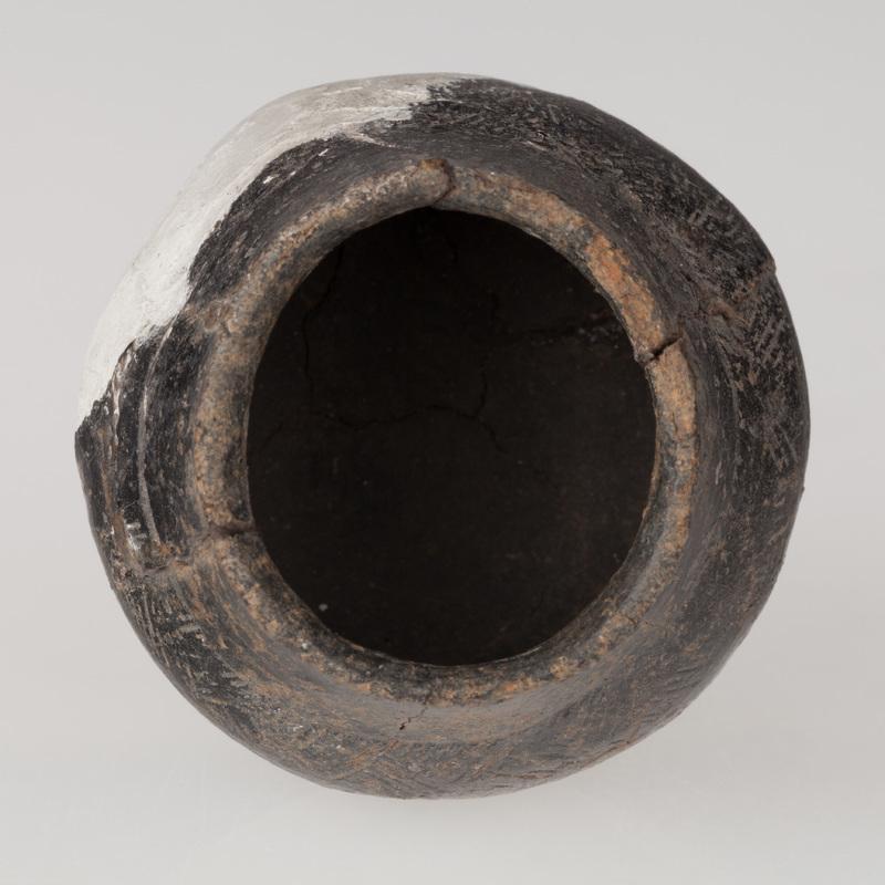 Miniaturowa waza o płaskim dnie, brzuścu baniastym i stożkowatej szyjce. Naczynie zdobione dookolnym żłobkiem i naprzemiennymi grupami kresek.